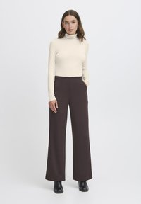 Sieviete stāv ar rokām kabatās, ģērbusies bēšā turtleneck, tumšās platajās biksēs un melnās potītes zābakos uz vienkārša balta fona.