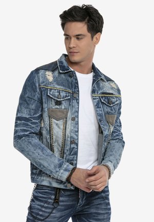 Jeansjacke in Hellblau mit abgeriebenen Stellen, silbernen Nieten, zwei Brusttaschen und Knopfverschluss. Über einem weißen T-Shirt getragen.