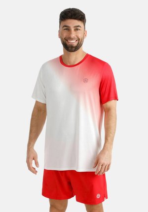 Mann lächelnd, trägt ein Sportshirt in Rot-Weiß-Verlauf und rote Shorts, steht vor einem schlichten hellen Hintergrund.