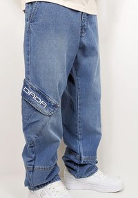 Blue denim cargo pants with a loose fit, featuring a large logo patch on the side pocket and subtle fading. Completed with white sneakers.