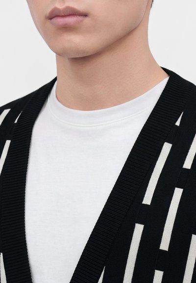 Gros plan sur le bas du visage et le cou d'une personne portant un t-shirt blanc à col rond sous un cardigan noir avec des motifs rectangulaires blancs verticaux.