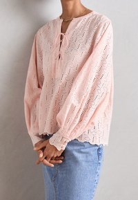 Femme portant un chemisier rose pâle en tissu ajouré avec des poignets boutonnés et un col à nouer, associé à un jean bleu clair, sur un fond uni.