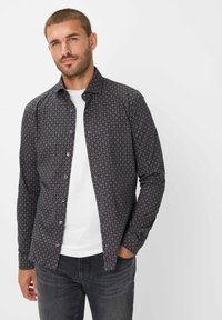 Camicia button-up in tessuto scuro con un motivo geometrico in rosso e beige. Maniche lunghe, colletto a punta, e abbinata a una t-shirt bianca sottostante.