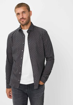 Button-up Hemd aus dunklem Stoff mit geometrischem Muster in Rot und Beige. Lange Ärmel, spitzes Kragen und kontrastiert mit einem weißen T-Shirt darunter.