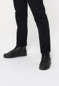 Czarne skórzane sneakersy w stylu high-top z zapięciem na sznurówki i teksturowanymi detalami. Płaska podeszwa i gładkie akcenty paneli. Noszone z czarnymi spodniami z mankietami.