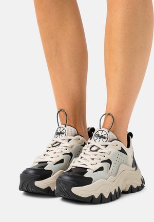 Baskets épaisses noires et blanches avec semelles épaisses et languettes, portées aux pieds sur un fond clair uni.