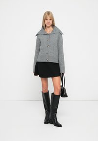 Gilet en tricot gris avec un grand col, cinq boutons et un ourlet côtelé ; associé à une jupe noire et des bottes noires hautes.