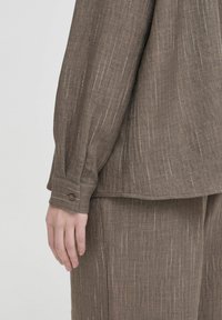 Vêtement en tissu texturé brun avec des rayures verticales. La manche présente un détail de bouton et une coupe décontractée, mettant en avant un design casual.