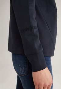 Personne portant une chemise noire à manches longues avec un texte subtil sur la manche et un jean bleu avec un texte brodé sur la poche arrière.