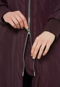 Langer burgunderfarbener Pufferjacke mit einem silbernen Reißverschluss, gerippten Bündchen und gestepptem Innenfutter. Der Fokus liegt auf dem teilweise geöffneten Reißverschluss.