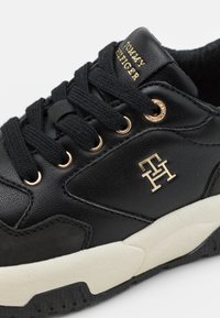 Zwarte sneaker van leer en stof, met gouden accenten, vetersluiting en een witte tussenzool. Logo aan de zijkant.