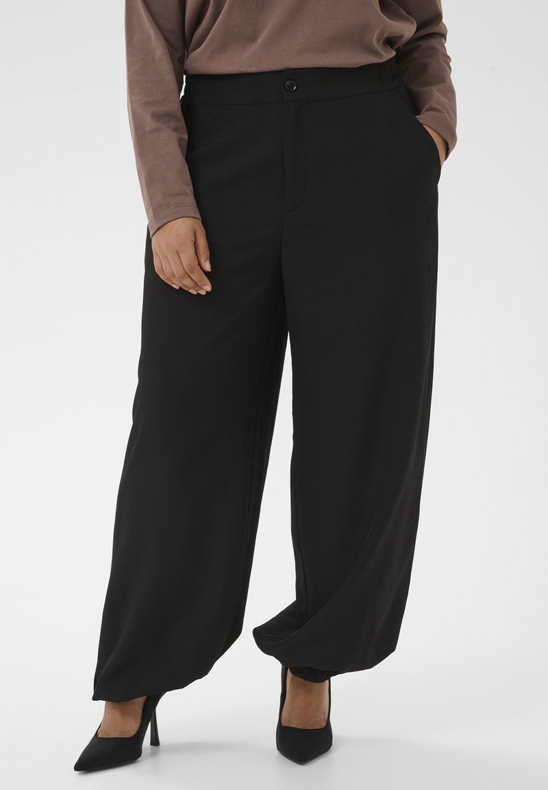 Personne portant un pantalon noir ample avec fermeture à boutons, une main dans la poche, associé à des chaussures à talons noirs et un haut marron à manches longues.