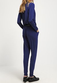 Mono azul marino con mangas largas, cintura elástica y bolsillos laterales. Textura de tela suave con un diseño de pierna cónica. Combinado con calzado negro.