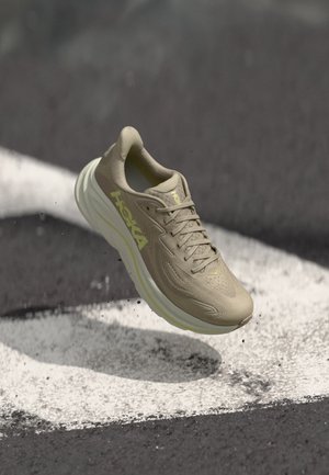 Chaussure de sport beige avec une tige respirante, un design texturé et un logo jaune proéminent. Elle est dotée d'une semelle rembourrée pour le confort.