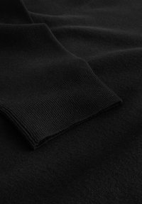 Crna pletiva s teksturiranim rebrastim manžetnama, prikazujući mekanu i glatku površinu s laganim sjajem. Fokus na čistim rubovima i šavovima.