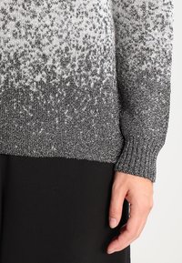 Ręka spoczywająca obok ciała, ubrana w melanżowy szaro-biały sweter z dzianiny z prążkowanym mankietem oraz czarne spodnie.