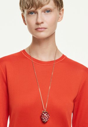 Persona con capelli biondi corti che indossa una maglietta rossa e una lunga collana d'oro con un grande ciondolo a forma di fragola rossa.