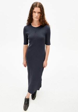 Frau mit langen lockigen Haaren, die ein tailliertes, marineblaues Midikleid mit Ellbogen-langen Ärmeln und schwarzen, mit Nieten besetzten Slip-On-Schuhen trägt.