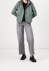 Grüne Windjacke mit Kapuze, schwarzes Oberteil, graue weite Jeans und schwarze Plateauschuhe. Die Windjacke hat seitliche Taschen und einen Reißverschluss.