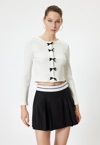 Top blanco de manga larga acanalada con cinco lazos negros en el frente, combinado con una falda negra con pliegues y una cinturilla de rayas blancas.