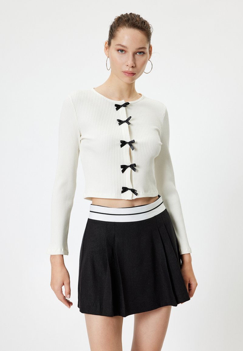 Top blanco de manga larga acanalada con cinco lazos negros en el frente, combinado con una falda negra con pliegues y una cinturilla de rayas blancas.