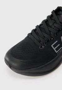 Black athletic shoe with a textured upper, rounded toe, lace-up design, and reflective accents on the side. Smooth rubber sole.