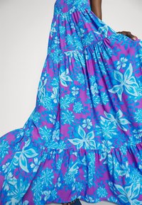 Robe maxi en tissu violet vif avec des motifs floraux bleus, dotée de volants en couches et d'une silhouette décontractée et fluide.