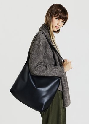 Sac en cuir noir à l'épaule avec une texture lisse, présentant un design doux et décontracté, et une seule sangle tenue par un mannequin portant un pull gris.