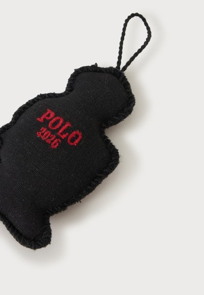 Μαύρο υφασμάτινο μπρελόκ σε σχήμα φιγούρας με κόκκινη κεντημένη επιγραφή "POLO 2026" και πλεγμένο λουράκι για κρέμασμα.