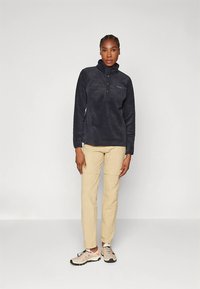 Flis pulover v temno sivi barvi z zadrgo na ovratniku, z žepom na prsih in belim logotipom. Kombiniran z bež hlačami in svetlimi čevlji.