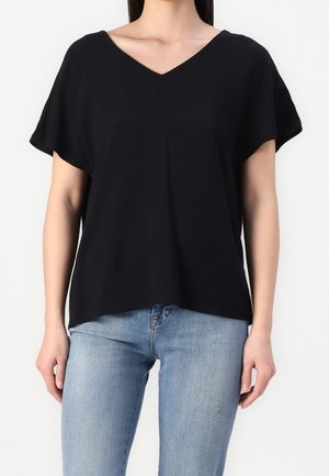 Femme portant un haut noir ample à manches courtes et col en V, associé à un jean ajusté bleu clair sur un fond uni.