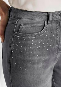 Gros plan sur un jean en denim gris avec une petite poche à pièces, décorée de clous en argent éparpillés près de la poche avant.