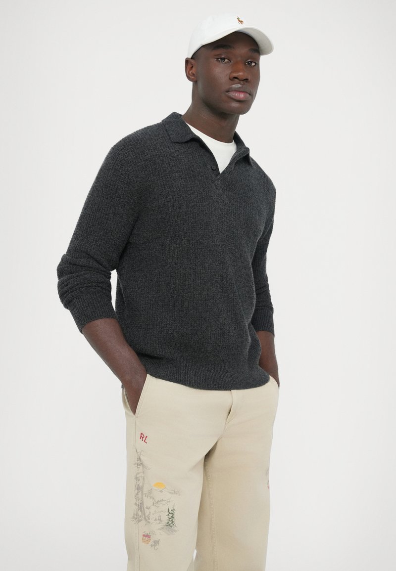 Maglione grigio scuro a maglia con colletto, indossato sopra una camicia bianca. Pantaloni beige con motivi ricamati all'aperto multicolore. Cappellino bianco.
