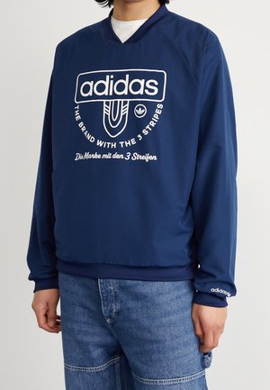 Osoba ubrana w granatową bluzę Adidas z białym logo i napisem, w połączeniu z niebieskimi dżinsami, stojąca na tle jednolitego jasnego tła.
