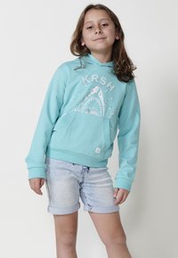 Tirkīza kapuci ar haizivs grafiku un "KRSH" tekstu, ar priekšējo kabatu un ribotām aprocēm. Pārī ar sarullētām džinsu šortiem.