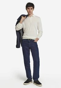 Vit pikétröja med lång ärm och krage, mörkblå jeans och svarta sneakers. En marinblå jacka hänger över ena axeln.