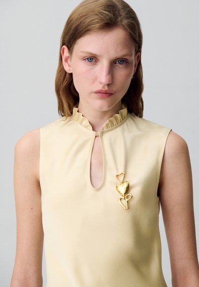 Haut sans manches beige avec un col froncé haut et une découpe en forme de clé. Présente des accents de broche en forme de cœur doré sur la gauche. Texture lisse.