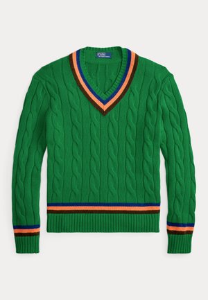 Homme portant un pull torsadé vert avec des rayures colorées, un pantalon en velours côtelé rose, des bottes vertes, des lunettes et une casquette marine avec le texte "Ralph".