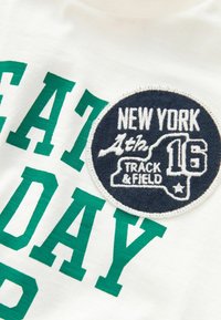 Hvid stof t-shirt med grønne blokbogstaver, der staver "EAT DAY." Har en rund marineblå lap med hvid tekst: "NEW YORK 16 TRACK & FIELD."