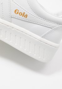 Sneaker bianco in materiale di pelle texturizzata, con suola in gomma dotata di scanalature verticali e logo "Gola" in rilievo color oro.