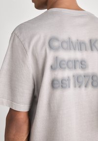 Šedé bavlněné tričko s vyraženým nápisem na zádech, který zní "Calvin Klein Jeans est 1978." Krátké rukávy a design s kulatým výstřihem.