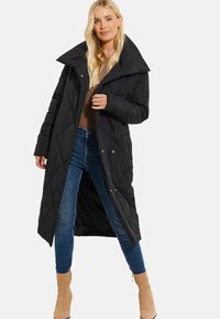Cappotto lungo trapuntato nero con un ampio colletto, bottoni a scatto e una linea dritta, abbinato a jeans blu e stivali alla caviglia beige.