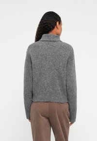 Kobieta z długimi kręconymi włosami, ubrana w szary sweter golf i brązowe spodnie, widoczna z tyłu na jednolitym tle.