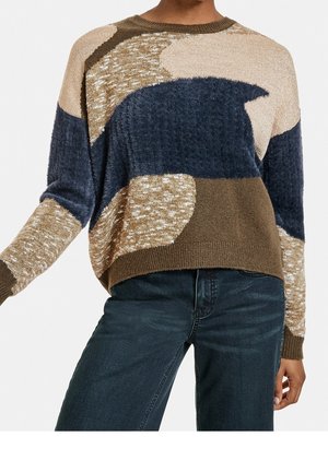 Kobieta nosząca sweter dziany o fakturze z abstrakcyjnymi blokami kolorów beżowego, brązowego, granatowego i kremowego, w połączeniu z ciemnoniebieskimi jeansami.