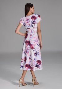 Floral midi-jurk met korte mouwen, witte stof met grote roze en paarse bloemen, taillering en een uitlopende rok. Gouden hoge hakken.