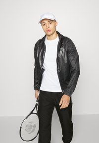 Svart hettejakke med strukturert overflate, hvit t-skjorte, svarte bukser og en hvit caps. Holder en tenniskølle med svart og hvite detaljer.