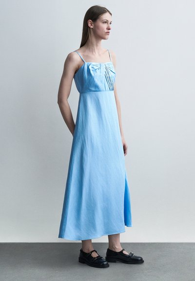 Femme en robe maxi sans manches bleu clair avec des détails plissés, debout sur un sol gris devant un mur gris clair uni.