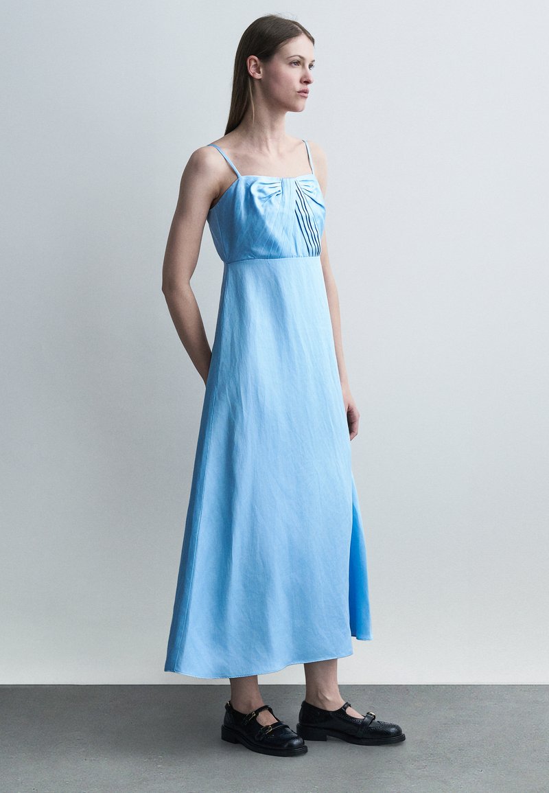 Γυναίκα με ανοιχτό μπλε αμάνικο μάξι φόρεμα με πιετέ λεπτομέρειες, στέκεται σε γκρι πάτωμα μπροστά από έναν απλό ανοιχτό γκρι τοίχο.