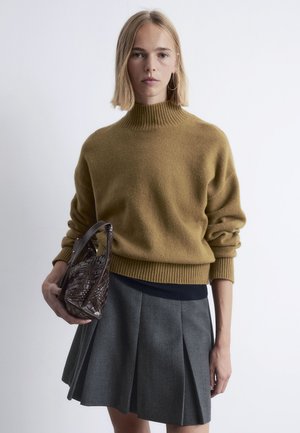 Mustard-colored knitted sweater with a high ribbed neckline, paired with a gray pleated skirt and a small brown woven handbag.