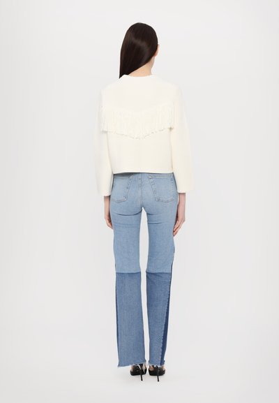 Weißer verkürzter Pullover mit Fransen am Rücken, kombiniert mit hellblauen Jeans aus Denim mit unterschiedlichen Farbtönen und Used-Effekten.